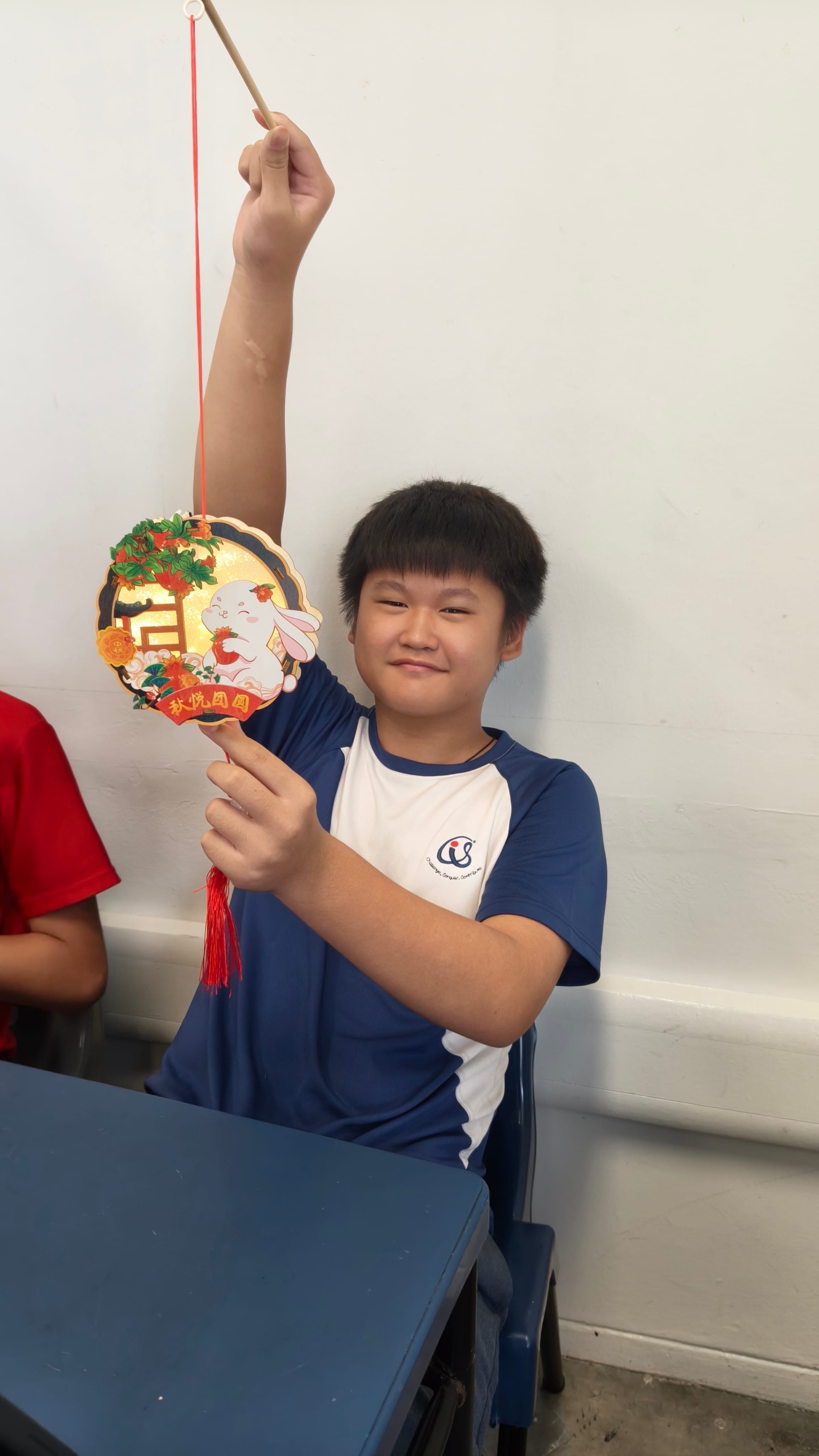 A male student in a blue-and-white PE shirt sits at a classroom desk, grinning and holding up a completed Mid-Autumn Festival lantern on a stick. The circular lantern features a white rabbit design with Chinese characters reading "秋悦圆圆" and a red tassel.